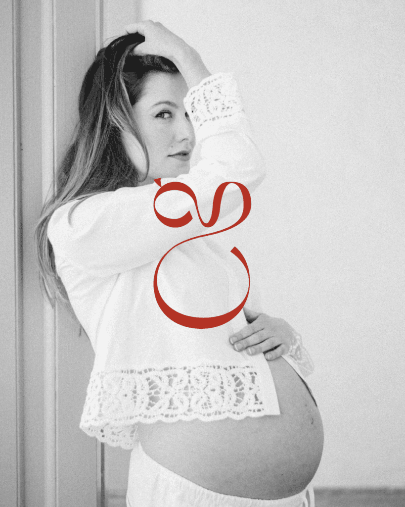 Black and white maternity portrait of a woman in a lace-trimmed blouse, tenderly cradling her baby bump with Christine Grace’s red submark logo overlaid.