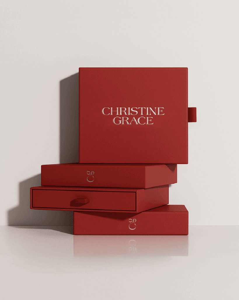 Stack of red branded boxes with “Christine Grace” elegantly printed on the top, styled on a light neutral surface.