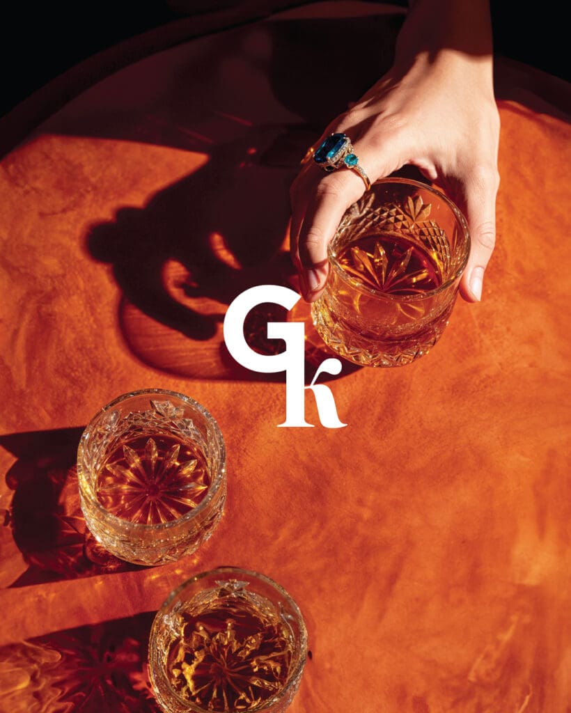 Close-up of hand with gemstone ring holding a cocktail on a warm-toned table, overlaid with Gk logo—capturing the brand’s mix of edge and elegance.